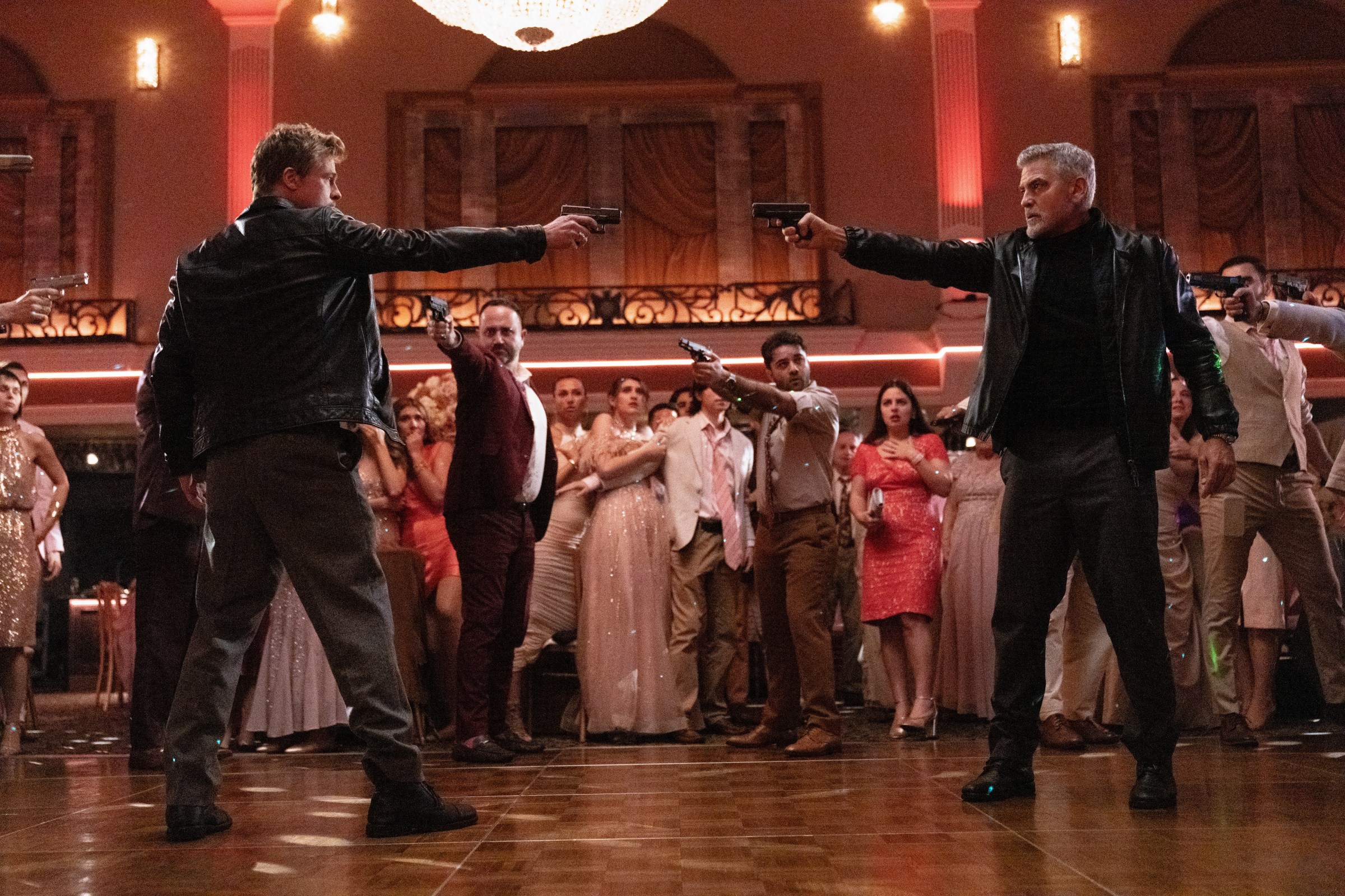 Brad Pitt and George Clooney point guns at each other, while other people point guns at them on a dance floor in Wolfs.