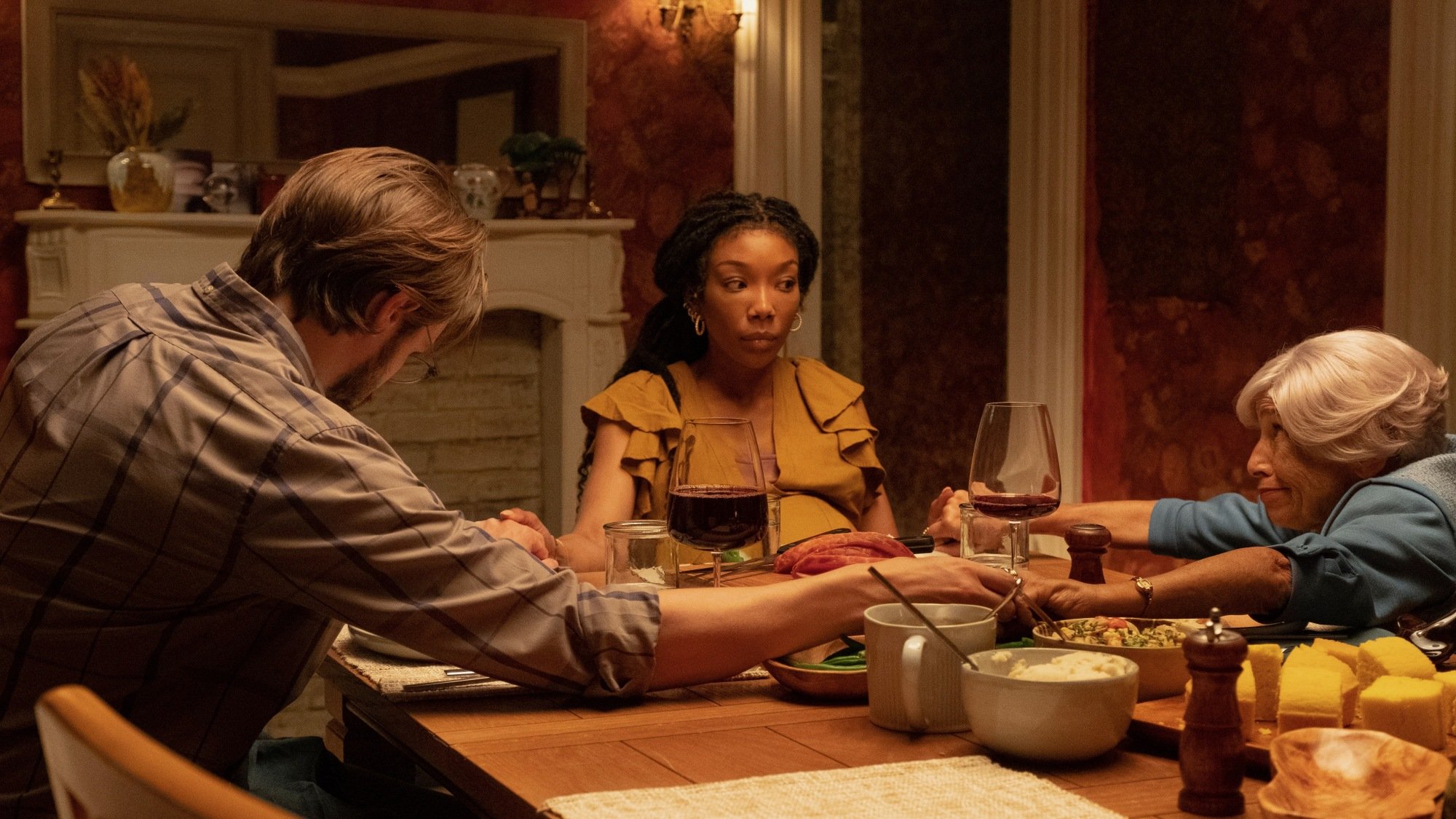 Brandy Norwood sits at a table, holding hands with a bearded man and an older woman in The Front Room.