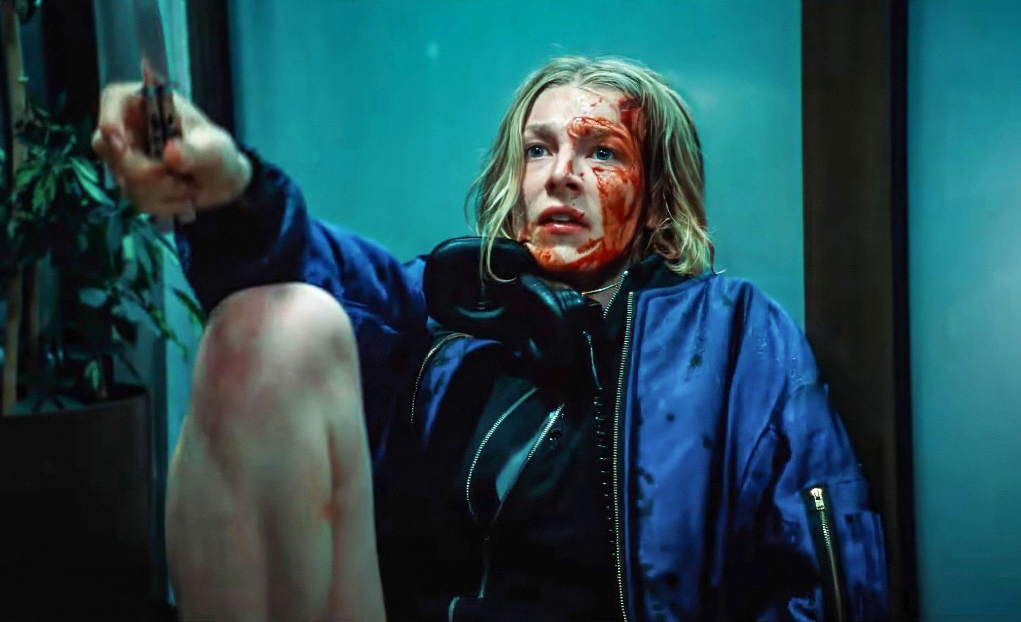 Gretchen (Hunter Schafer from Euphoria), a teenage girl with her face and hands covered in blood, sits outside against a glass door and waves a knife at an unseen assailant in Tilman Singer's Cuckoo