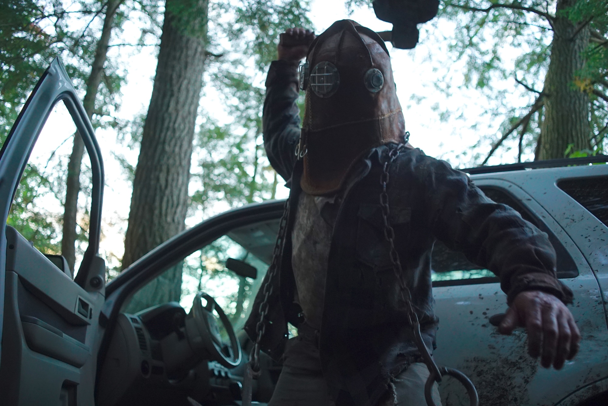 A hooded figure standing in front of a dirty white four-door bus raises an axe to strike. They have chains draped around their shoulders and appear to be standing in the middle of the forest. (In a Violent Nature)