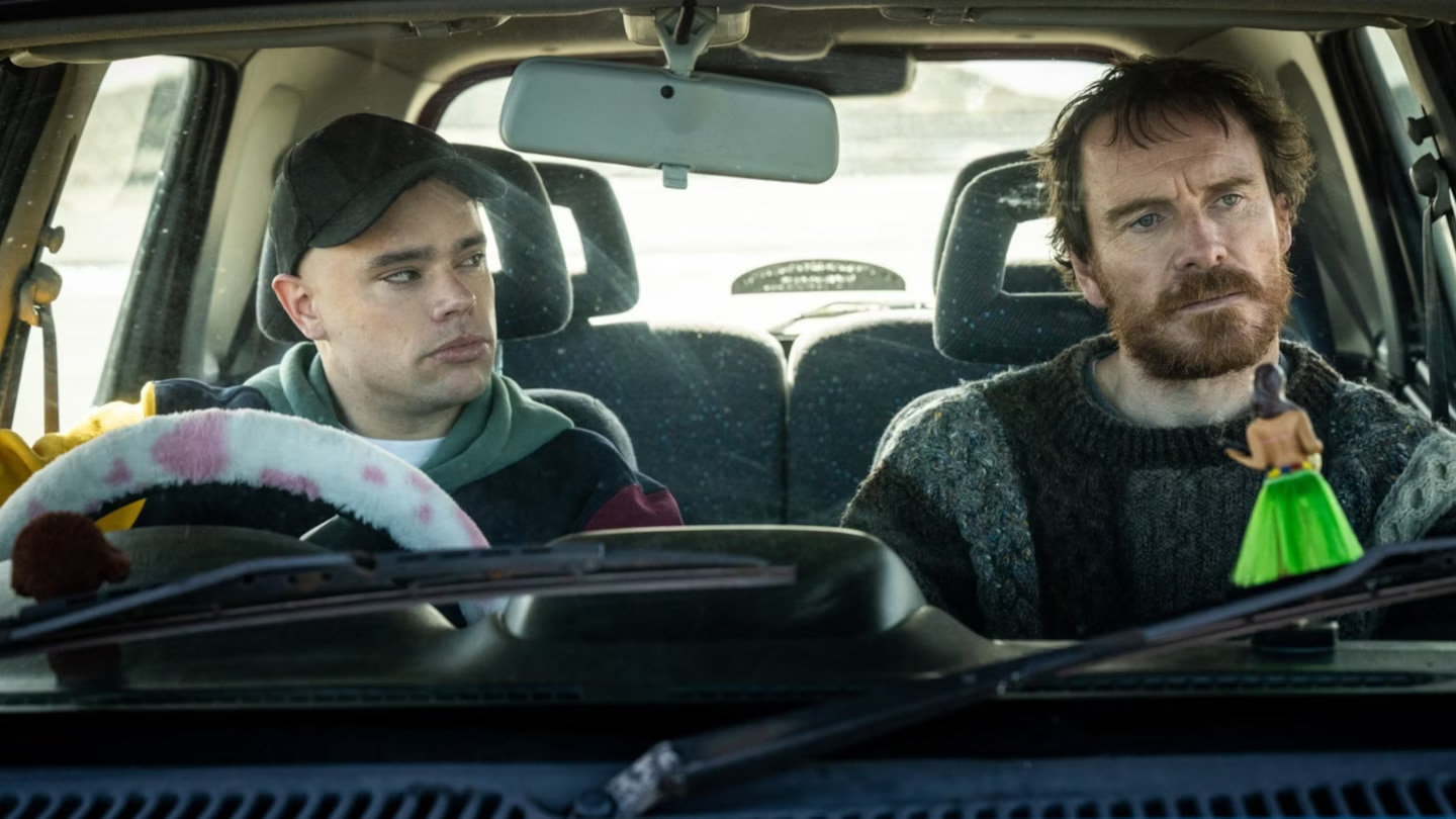 Michael Fassbender and a man wearing a black baseball cap sit in a car in Kneecap.