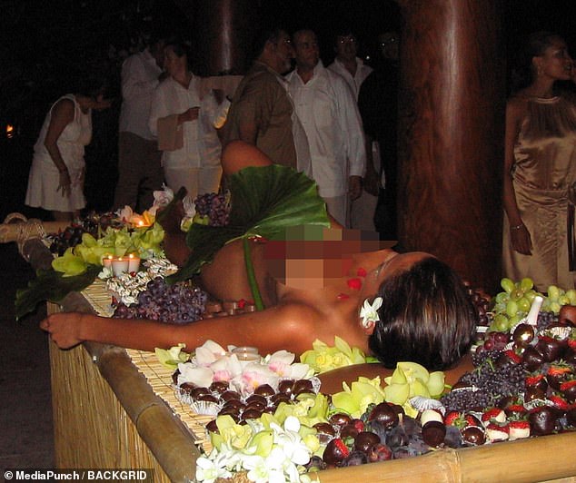 The woman on the table is surrounded by fruit for the guests, and a single green leaf strategically placed to cover her genitals