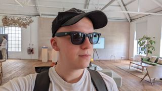 Hamish is wearing black Wayfarer smart glasses by Ray-Ban and Meta. He is also carrying a hat and a bag in a large modern living room.