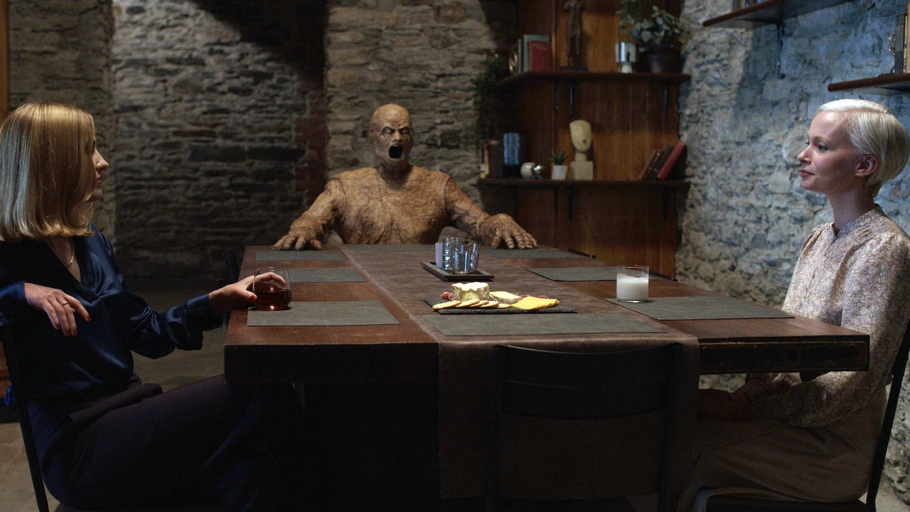 Two women sit across from each other at a table with a creepy wooden figure in Oddity.