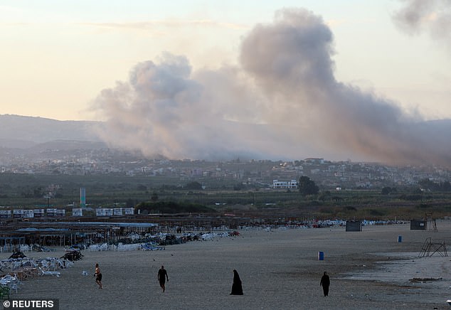 People walk on a beach as smoke rises over southern Lebanon after Israeli attacks