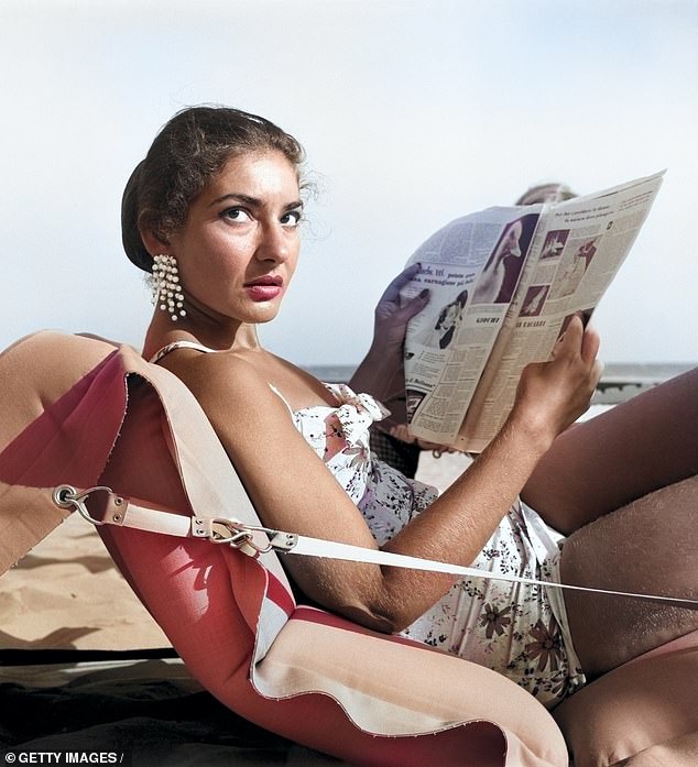 American-born Greek opera singer Maria Callas lies on an air mattress on the beach at Venice Lido, wearing a floral swimsuit and dangling earrings, reading a newspaper, Venice, 1950