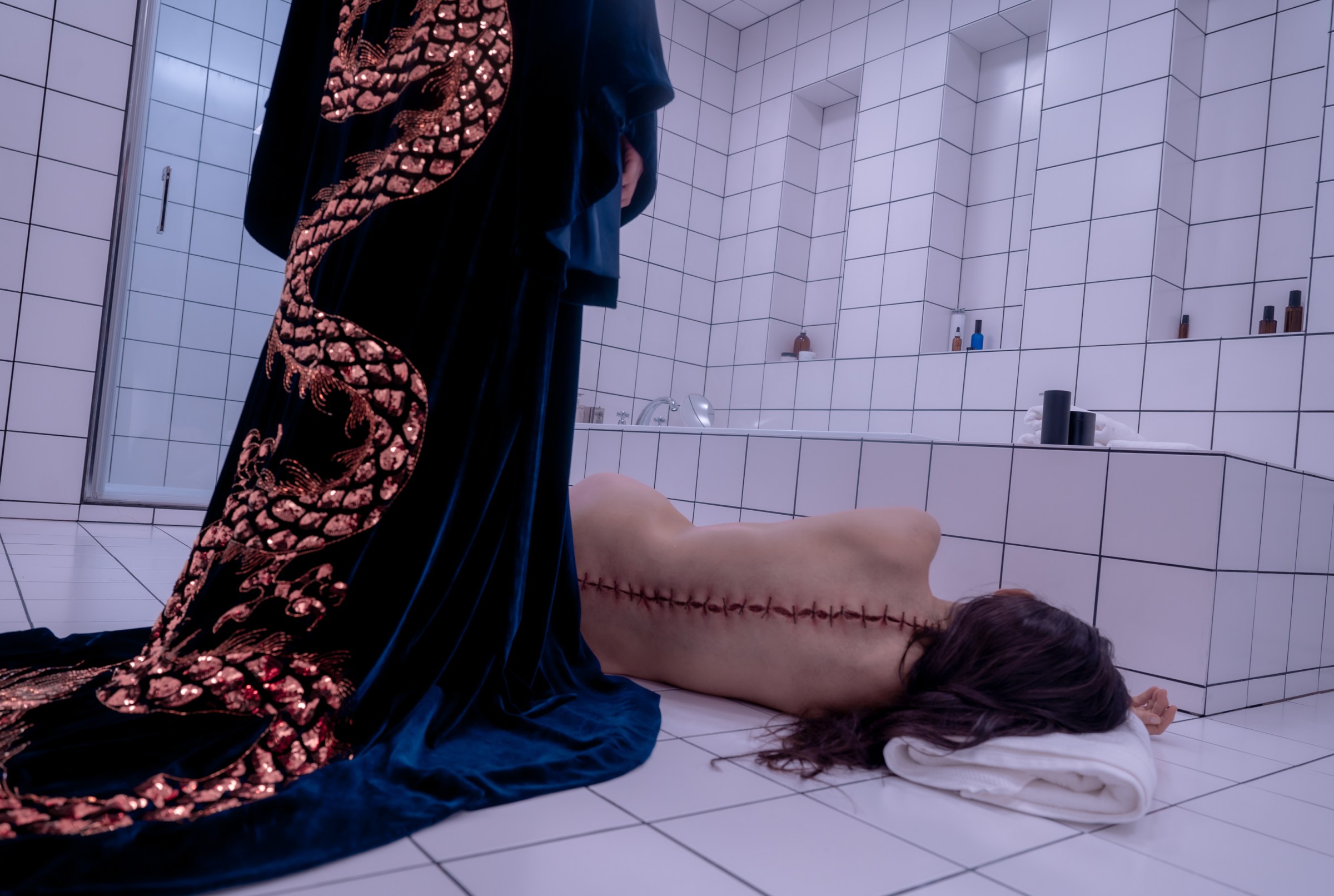 Sue looks at Elisabeth's body on the bathroom floor, with a giant incision scar on his back