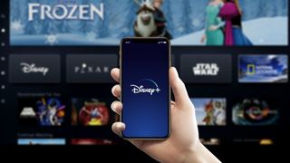 Disney Plus logo on a smartphone on the right with the app loaded on a TV in the background