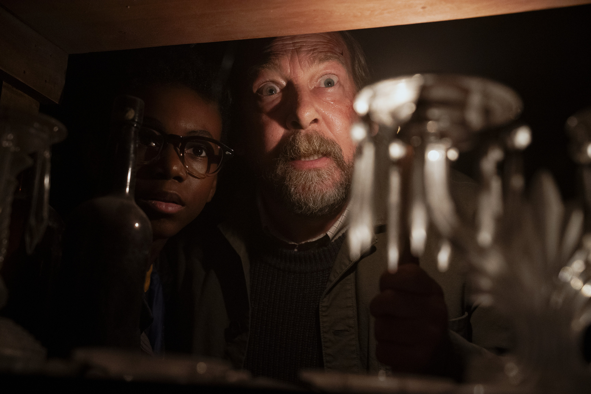 A bearded man and a teenager in glasses look through a shelf of glassware while illuminated with a flashlight in Salem's Lot  