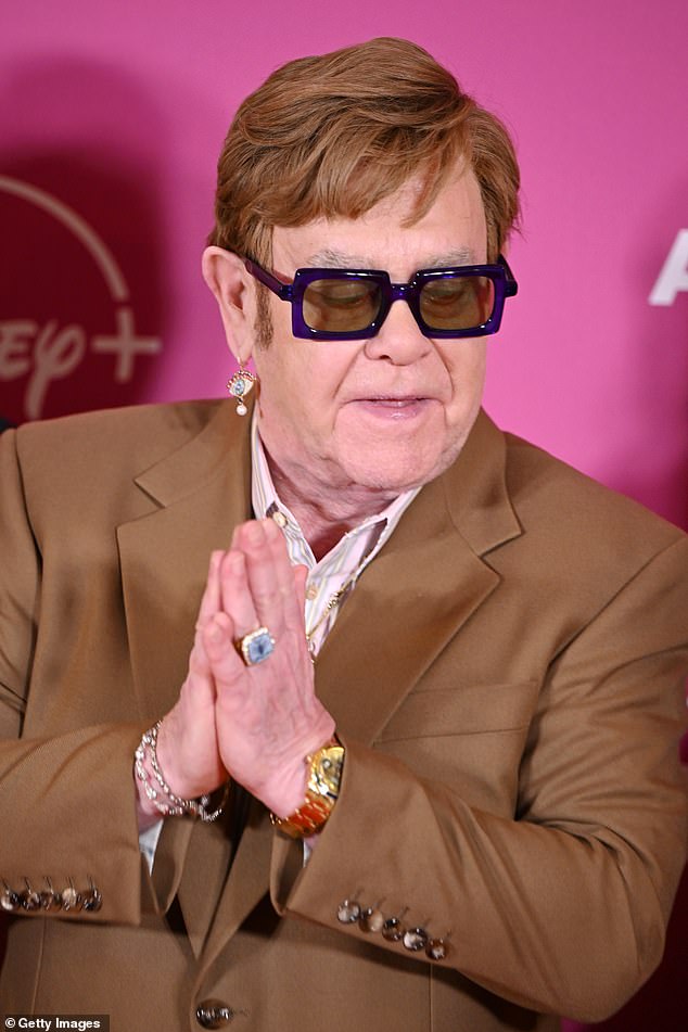 Looking as trendy as ever with his signature glasses, Elton wore a dark brown suit with a pink pinstripe shirt