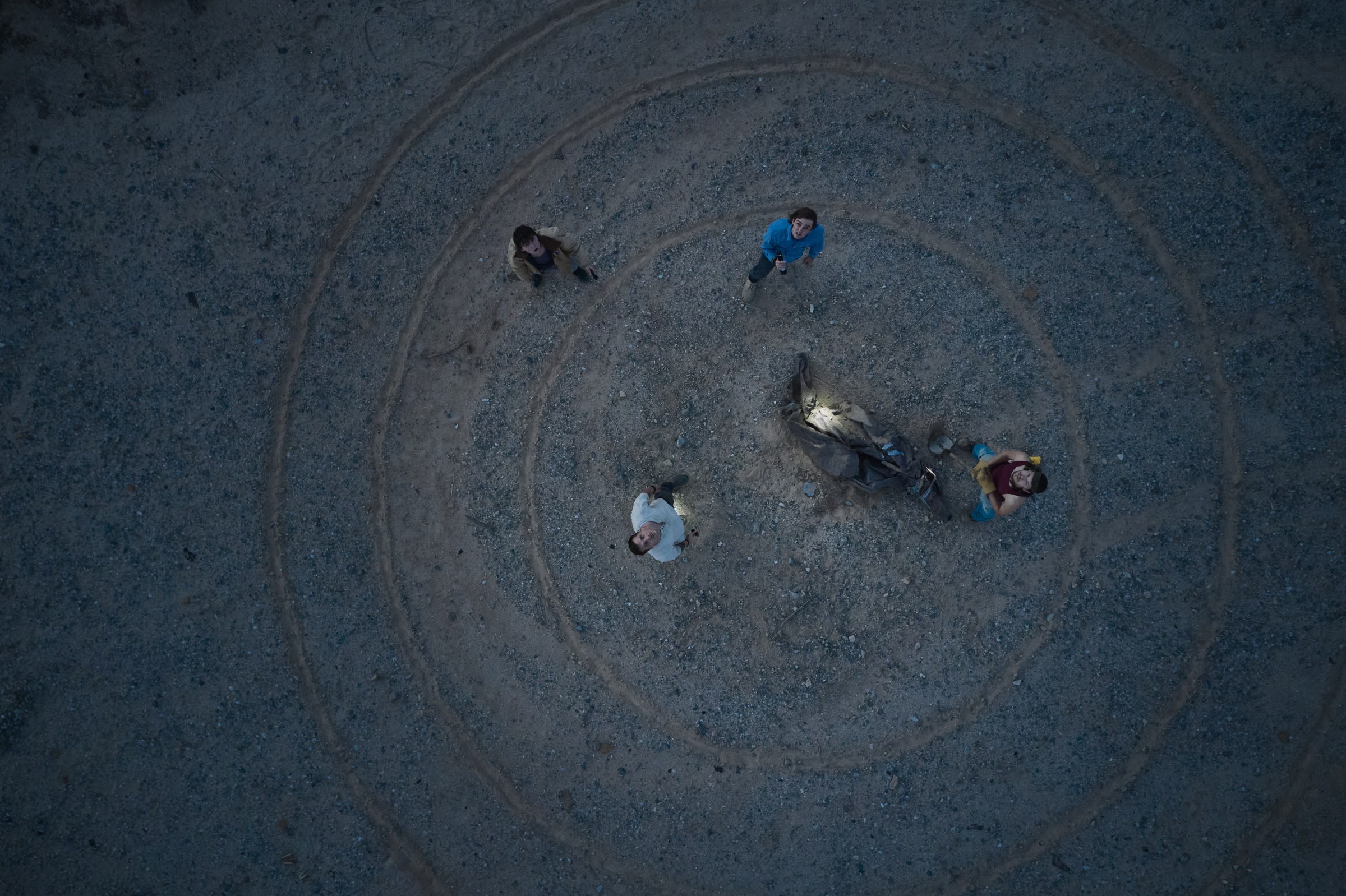 An overhead shot from Falling Stars of four young men standing around an exhumed body in the desert. They look up at the camera. They are surrounded by circular lines drawn in the dust.