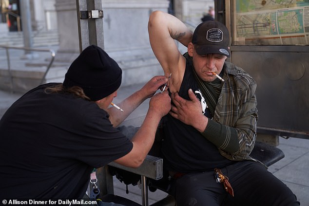 A homeless man injects fentanyl into his friend's armpit, due to a lack of usable veins, as people walk by near City Hall in San Francisco in September 2022