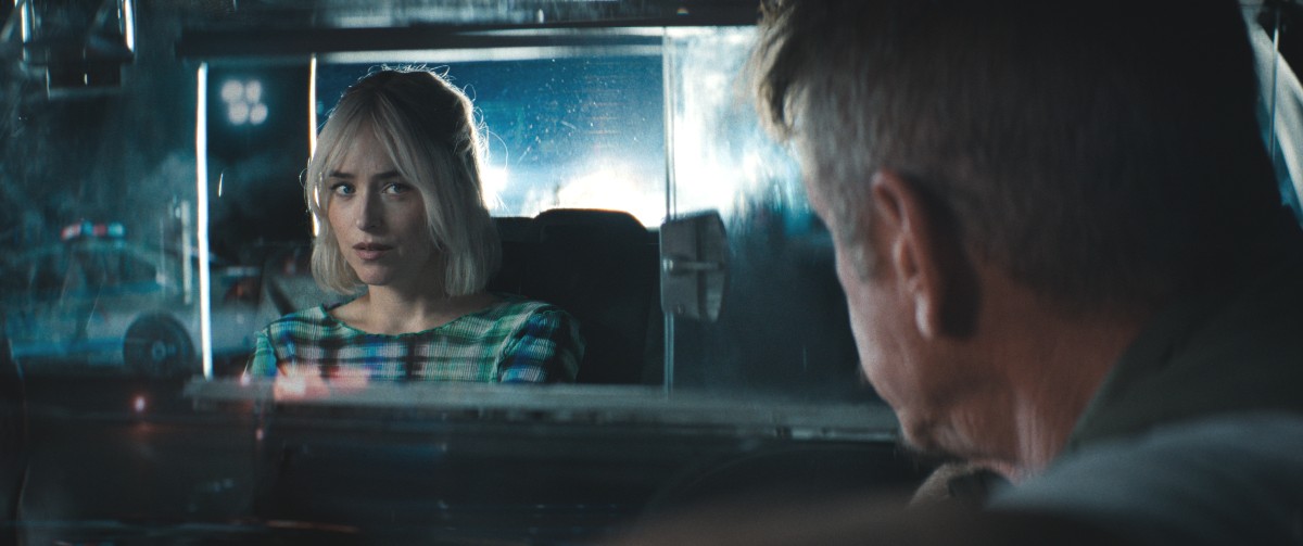 A male taxi driver looks at a female passenger with platinum blonde hair in the backseat of his taxi in Daddio.