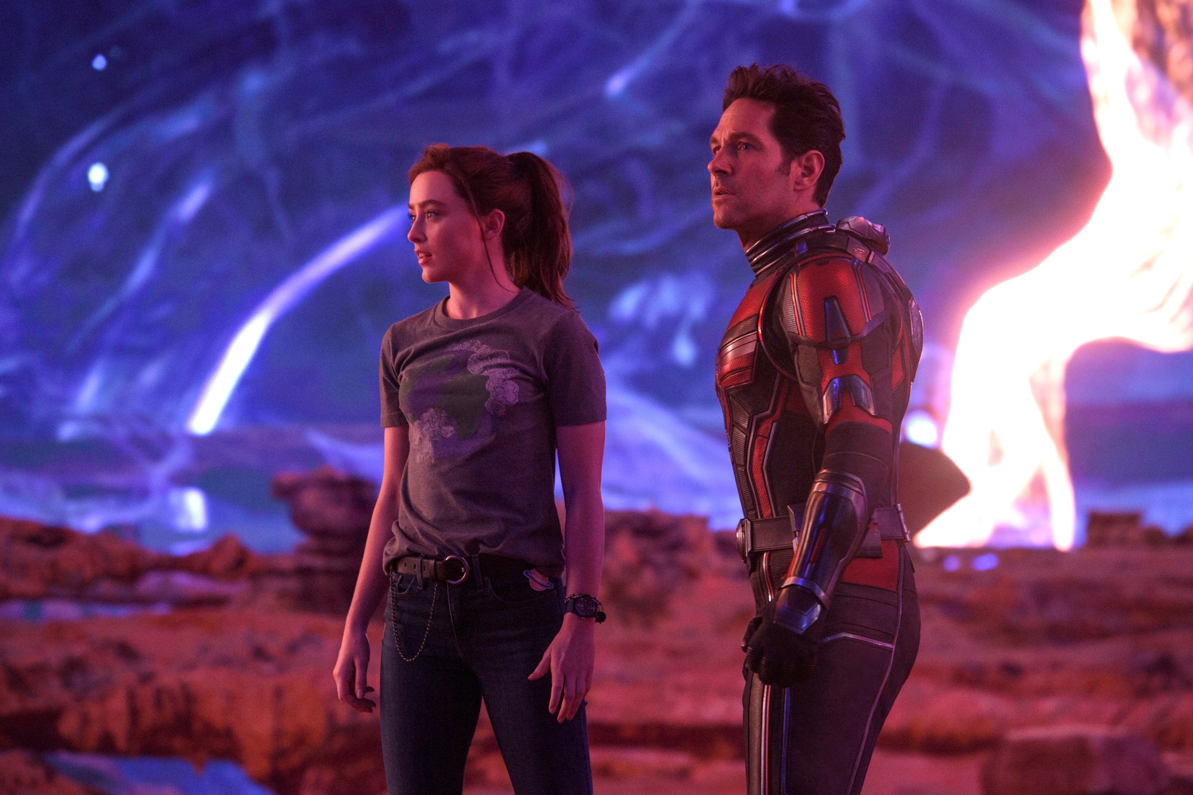 (L-R): Kathryn Newton as Cassandra “Cassie” Lang and Paul Rudd as Scott Lang/Ant-Man in Ant-Man and the Wasp: Quantumania. Cassie wears jeans and a T-shirt, while Scott wears his Ant-Man costume with the helmet off. They stand stunned at an otherworldly vista of swirling skies.