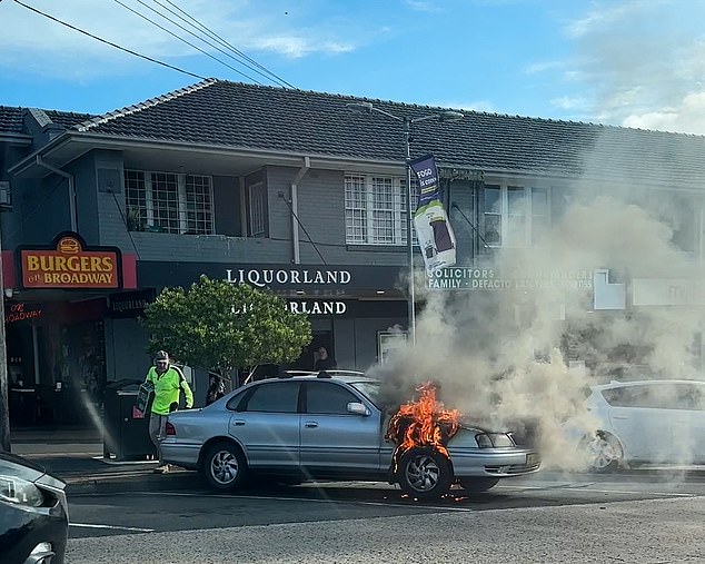 Surreal footage shows the bonnet of a car bursting into flames as black smoke pours from the vehicle parked outside a liquor store in the Sydney suburb of Ermington. The driver then emerges from the nearby store with a case of beer in hand