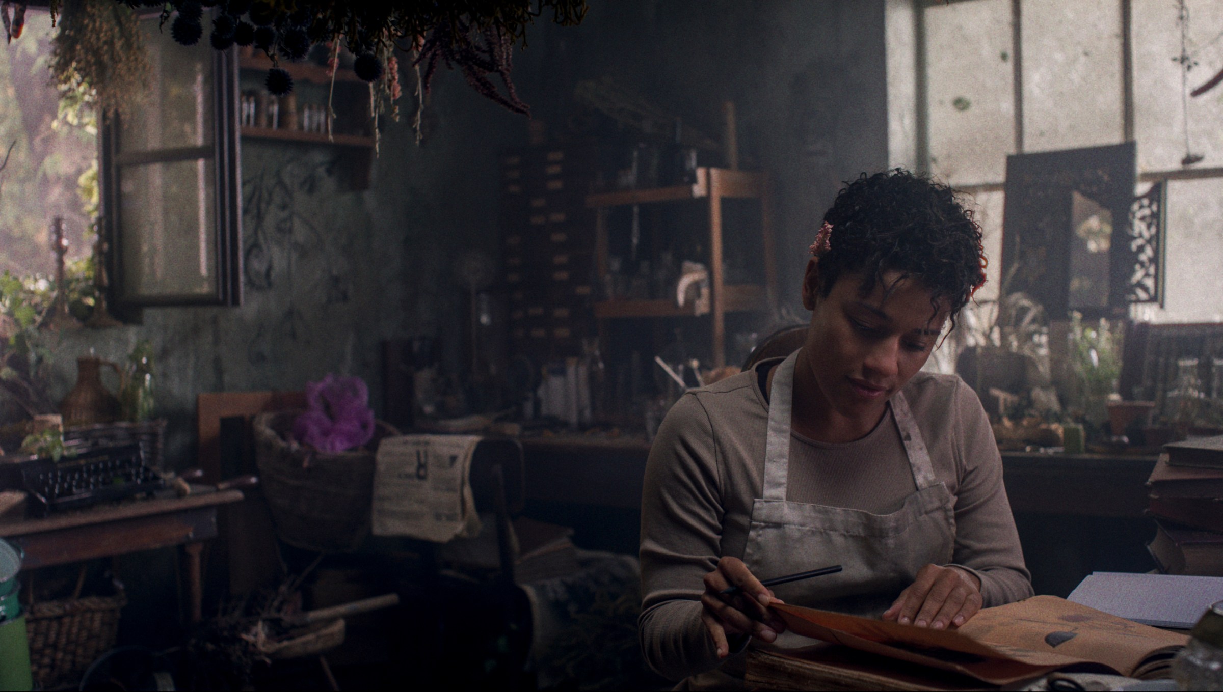 Chef (Ariana DeBose) examines sheets of parchment and paper in a dark, overgrown, cluttered mansion kitchen in House of Spoils