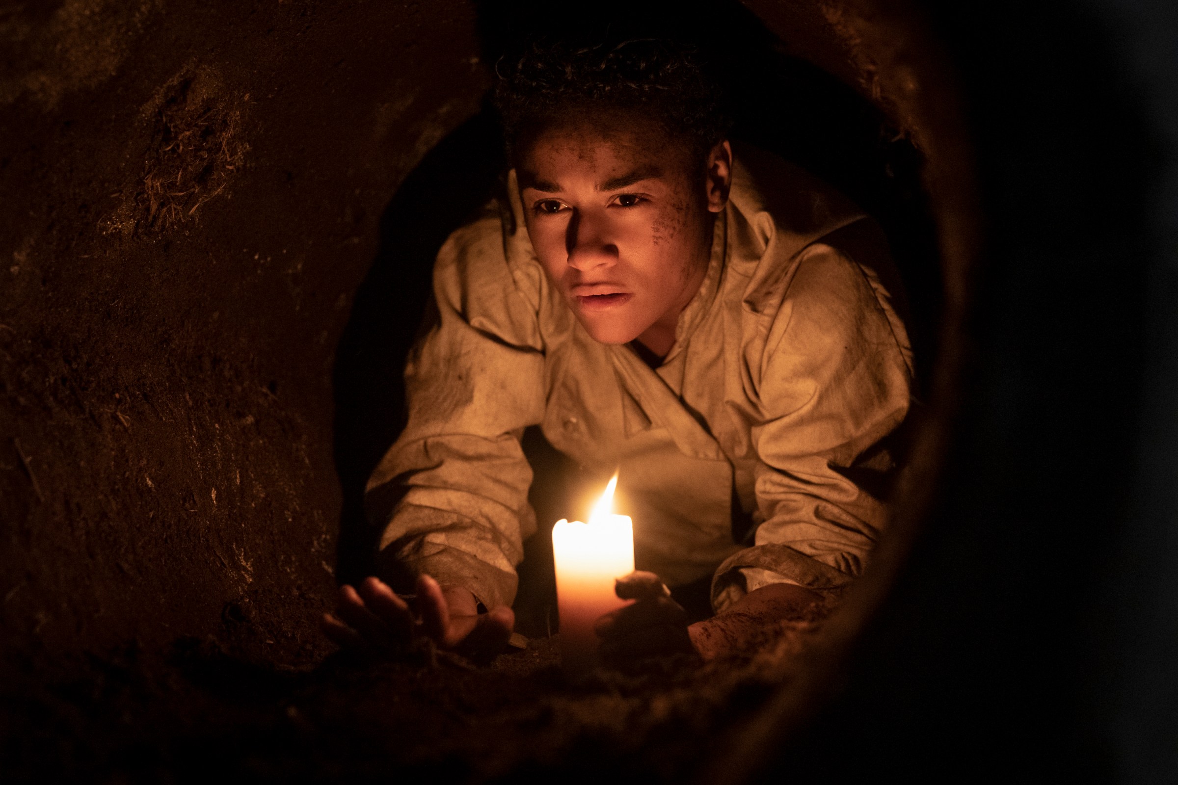 Chef (Ariana DeBose), candle in hand, crawls through a narrow underground tunnel in House of Spoils