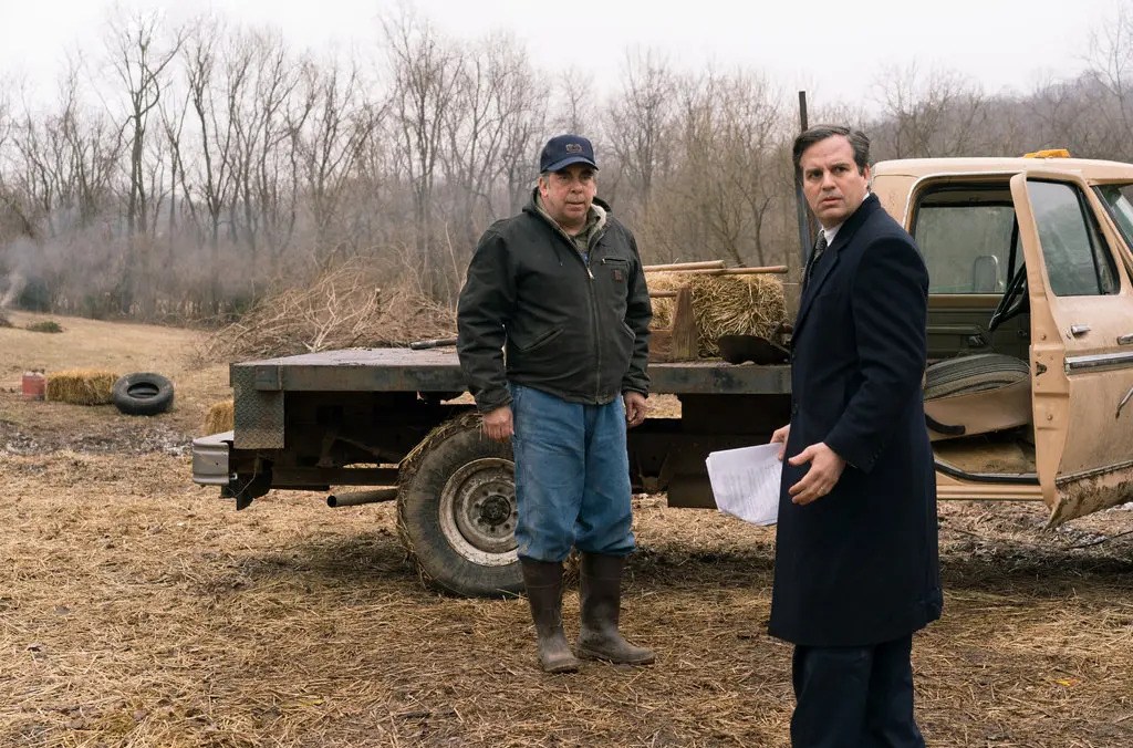 A man in a dark overcoat looks worriedly over his shoulder next to a man standing next to a flatbed truck in Dark Waters.