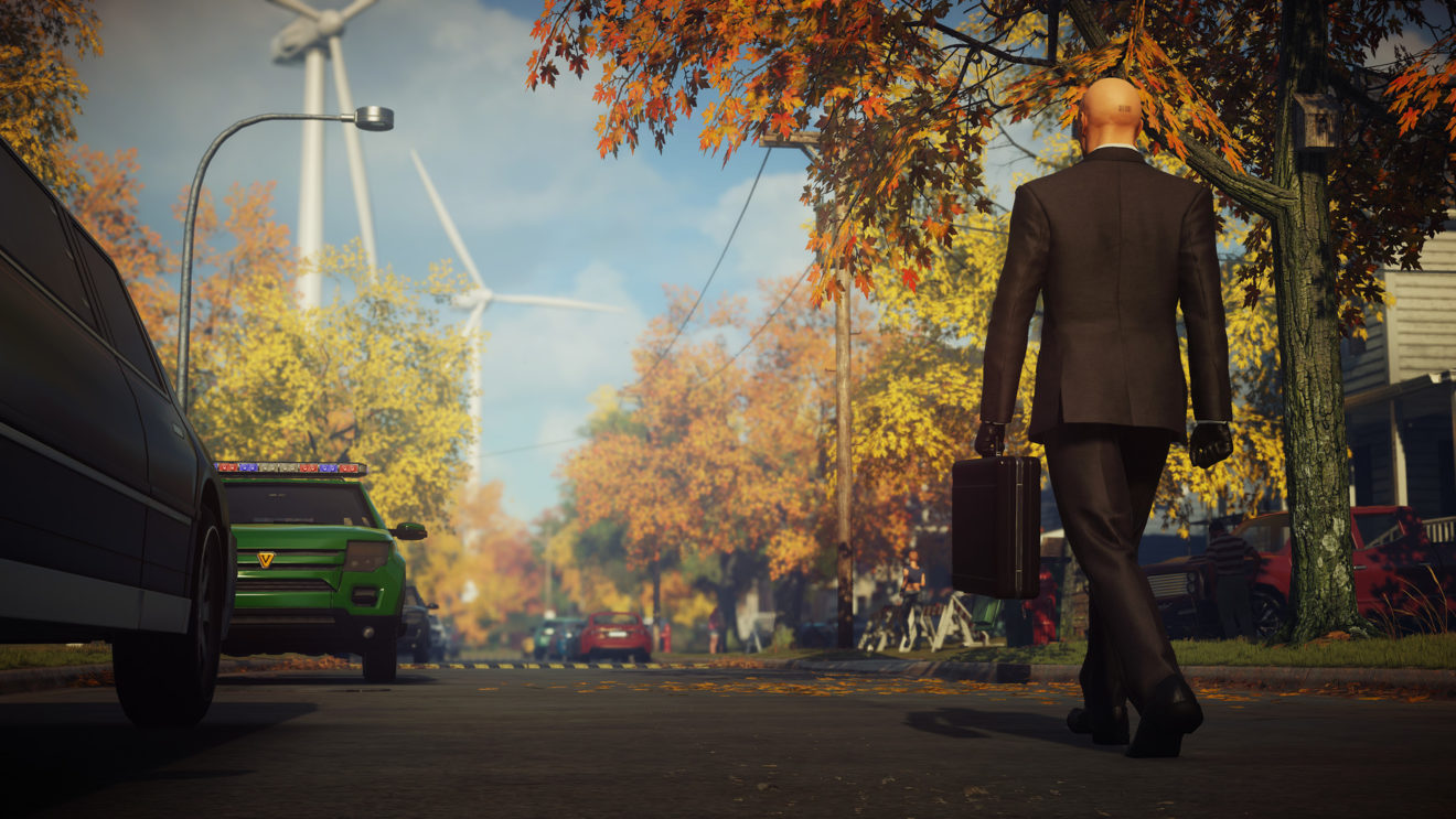 Agent 47 holds a briefcase as he walks down a suburban street with windmills visible in the distance in Hitman: World of Assassination.