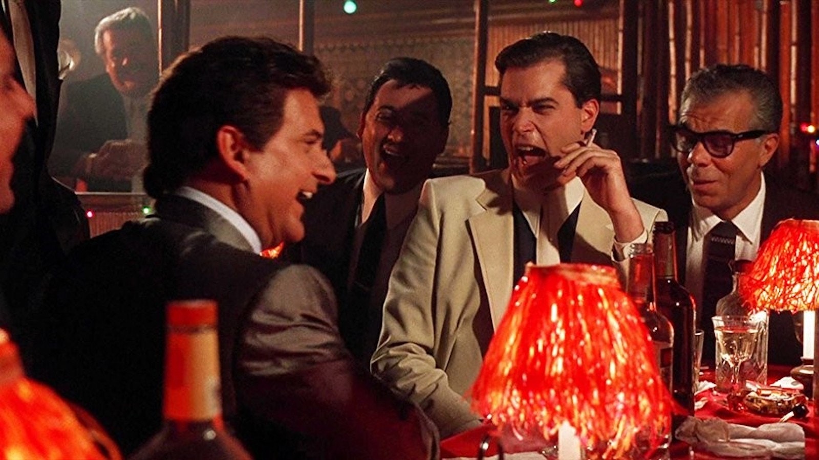 Joe Pesci and Ray Liotta laugh at a table surrounded by men in suits in Goodfellas.