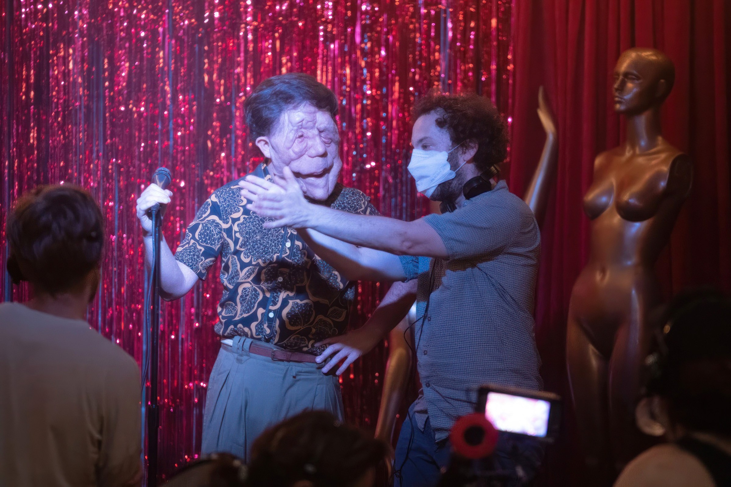 Actor Adam Pearson stands in front of a karaoke microphone while director Aaron Schimberg, wearing a white surgical mask, holds his hands up on the set of A Different Man