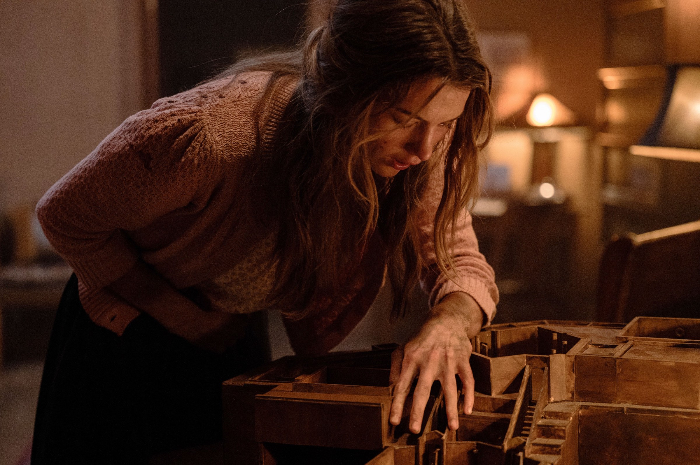 Nurse Paxton (Chloe East, wearing a pink sweater, with her hair dangling around her face) clutches her stomach as she leans over a small wooden model of the house she is trapped in in the horror movie Heretic