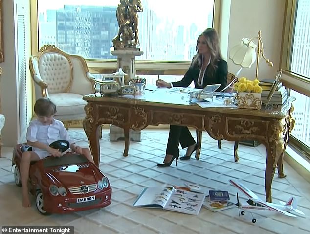 Melania sits at an ornate gold desk in a sharp black suit while her son plays in his toy car