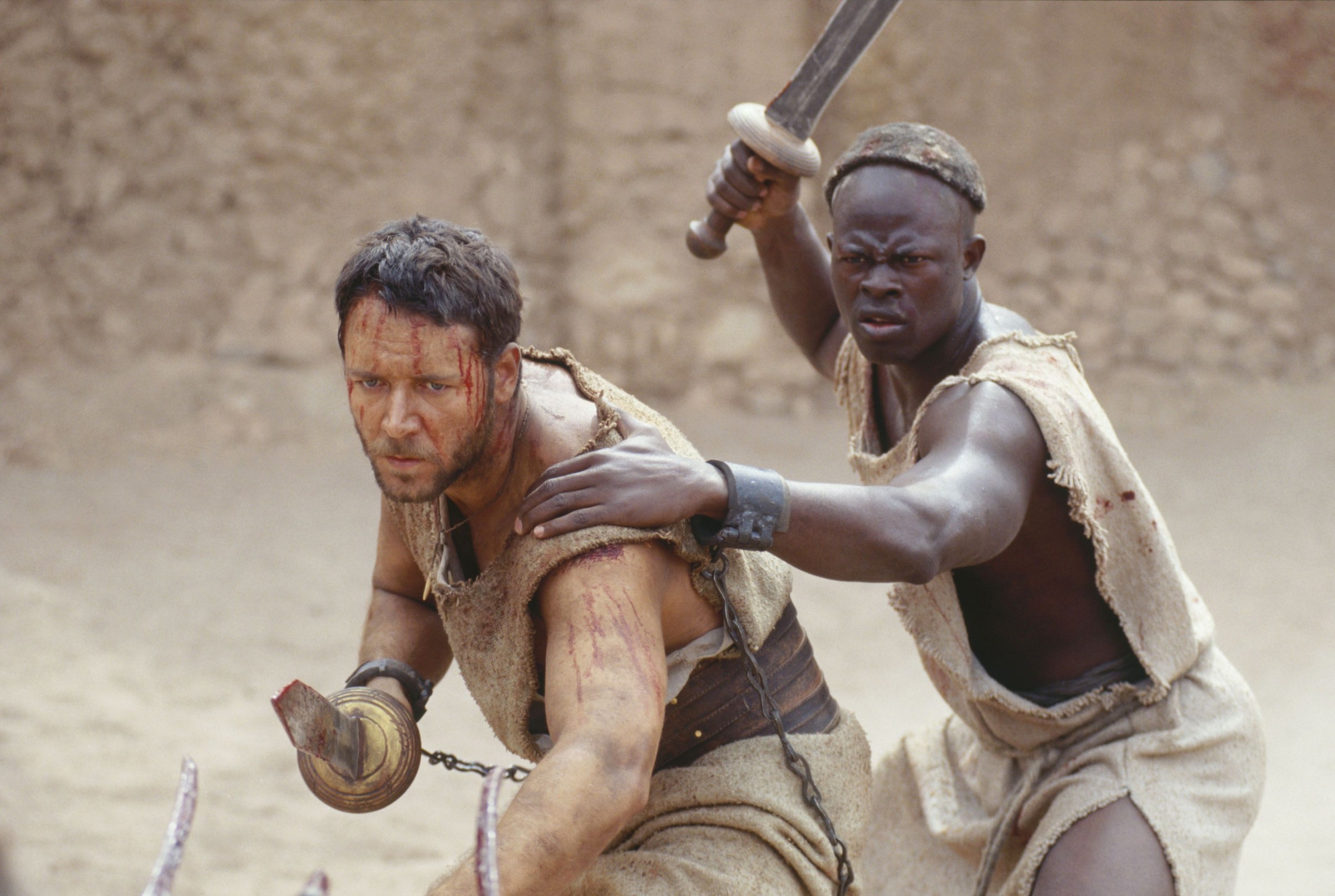 A pair of bloodied, dusty gladiators (Russell Crowe, Djimon Hounsou) stand together in the Colosseum, swords at the ready, in 2000's Gladiator