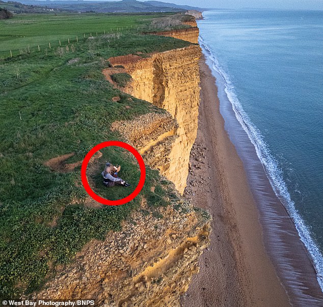 The two unidentified women were seen arm in arm as they sat uncomfortably close to the dangerous cliff edge at West Bay to watch the sunset (pictured)