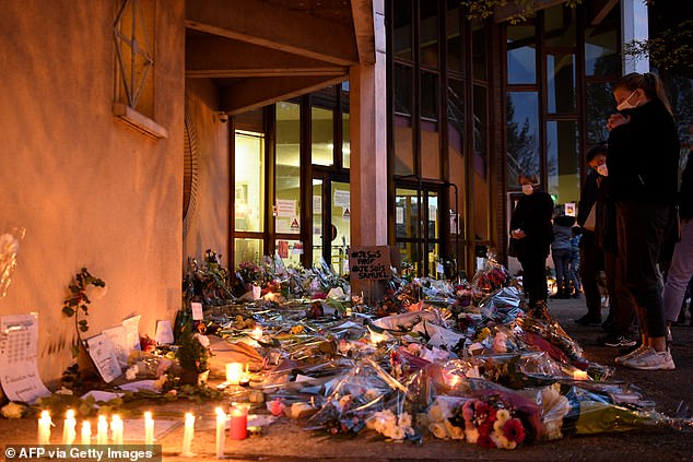 People stand in front of flowers and candles next to a sign reading 