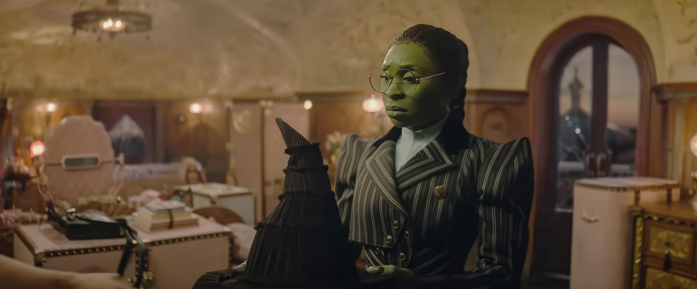 Elphaba (Cynthia Erivo), in a pinstripe suit with dramatic shoulder pads, looks sadly at the pointy black witch hat her supposed friend Glinda gave her in Wicked: Part I