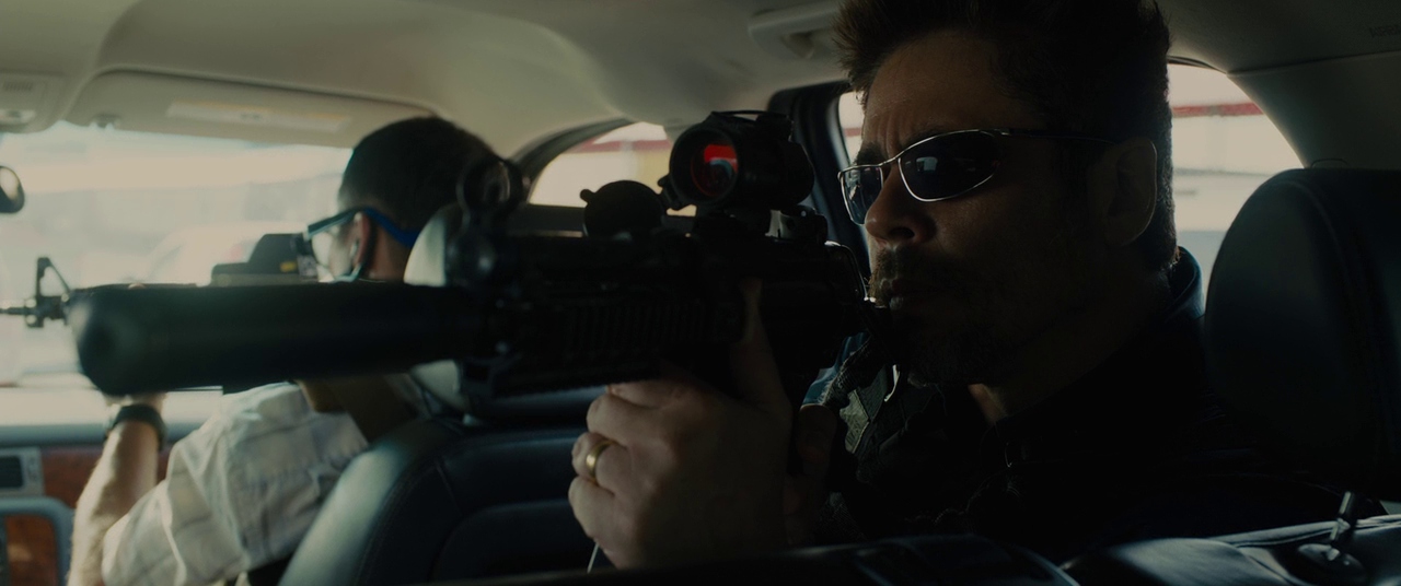 Benicio del Toro wears sunglasses and holds a silenced rifle in a car in Sicario.