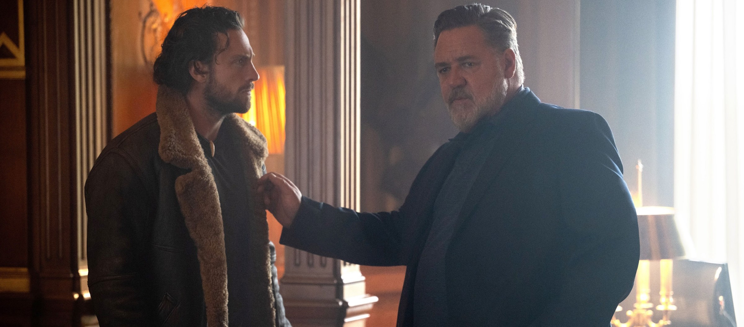 Russian gangster Nikolai Kravinoff (Russell Crowe, in a dark suit) stands with his hand on the shoulder of his extremely large adult son Sergei (Aaron Taylor-Johnson) in Kraven the Hunter