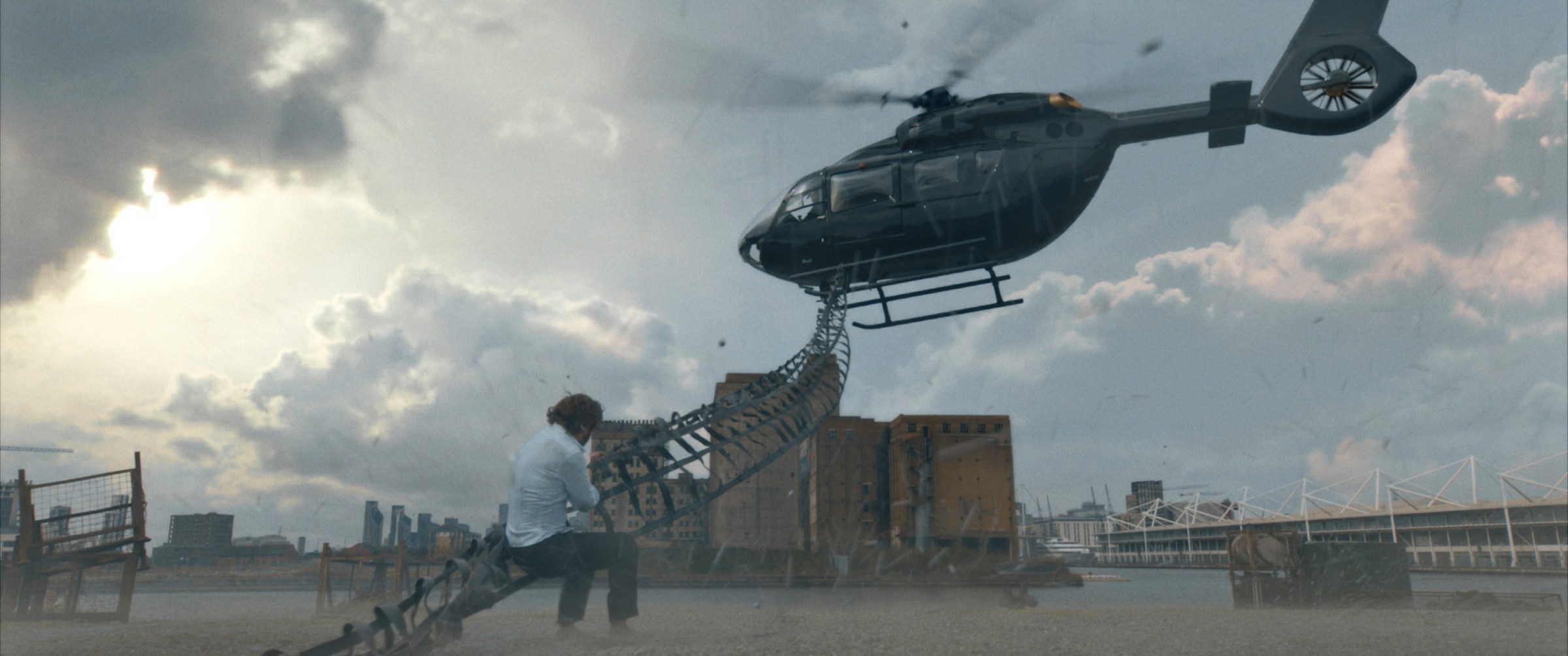 Kraven (Aaron Taylor-Johnson) stands in a dusty field surrounded by disused buildings, hanging from a strip of heavy canvas attached to the skids of a helicopter flying above in a very digital-looking action scene from Kraven the Hunter