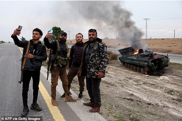 Armed men pose for photos near a military vehicle belonging to Syrian regime forces and seized by anti-government forces, which is on fire after being struck by regime forces, in Hama Governorate, December 7 2024