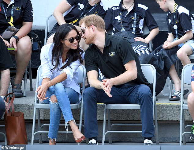 The prince whispers to Meghan, who is wearing Misha Nonoo's The Husband shirt