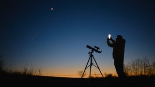 A man standing next to a telescope and holding his smartphone up to the moon and the night sky