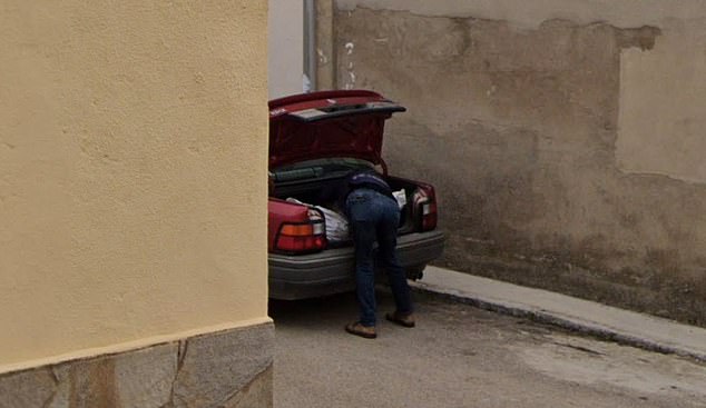 The photo shows a man loading what appears to be a body bag into the trunk of a red car