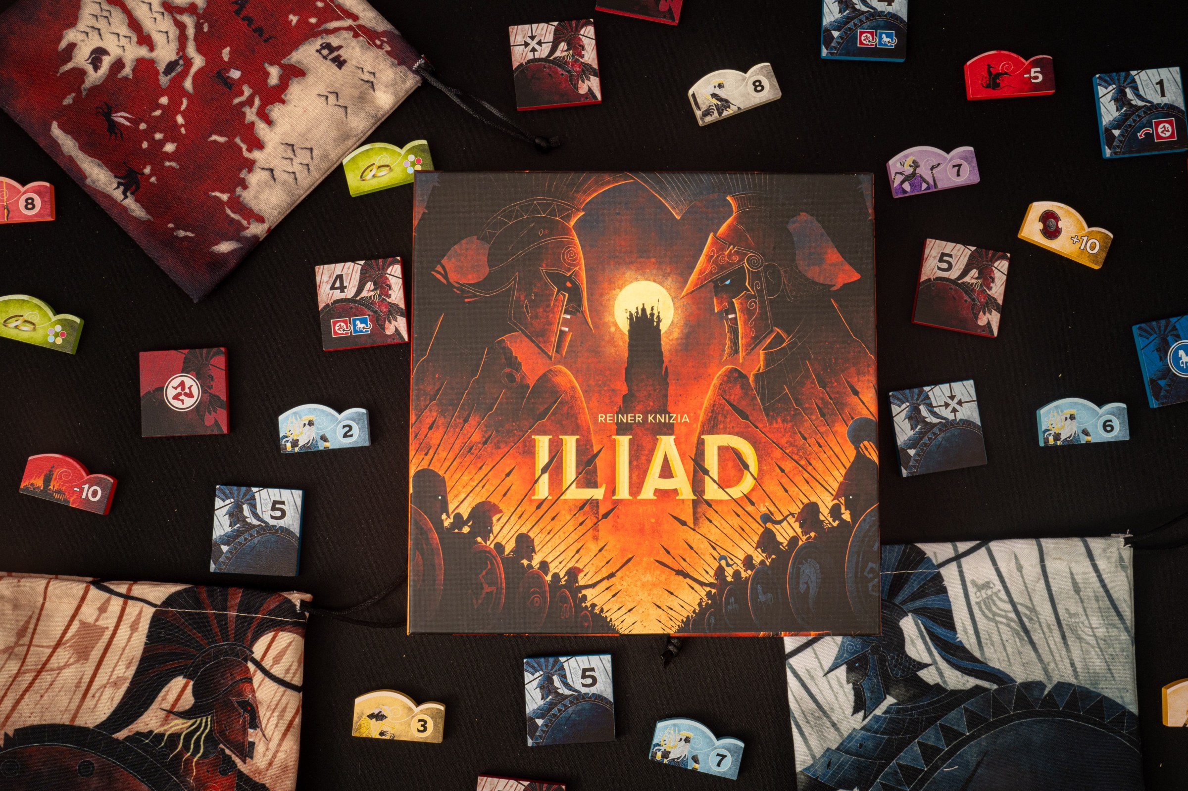 The box for Iliad sitting on a table, surrounded by colorful tokens depicting gods and Greek warriors.