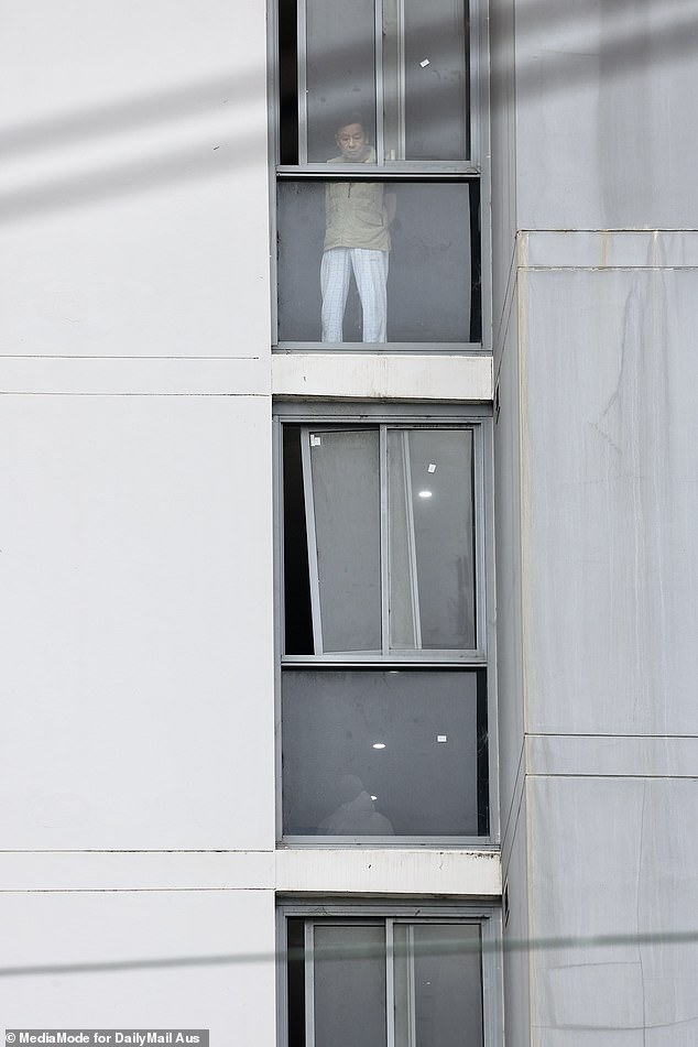 A resident is seen peering out the window of his unit as emergency services arrive