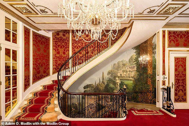 The opulent mansion features a spiral staircase, covered in thick dark red carpeting, a formal drawing room with a grand Italian crystal chandelier, surrounded by gilded custom paneling and ceiling heights of over 10 feet.