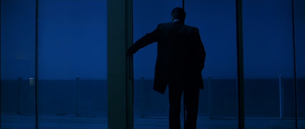 A man in a suit stands next to a window overlooking a beach at night in Heat.