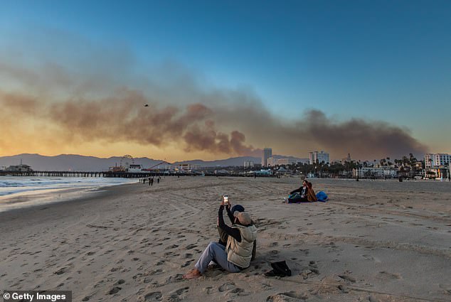 A woman takes a photo on the beach with the Santa Monica Pier in the background with smoke from the Palisades Fire