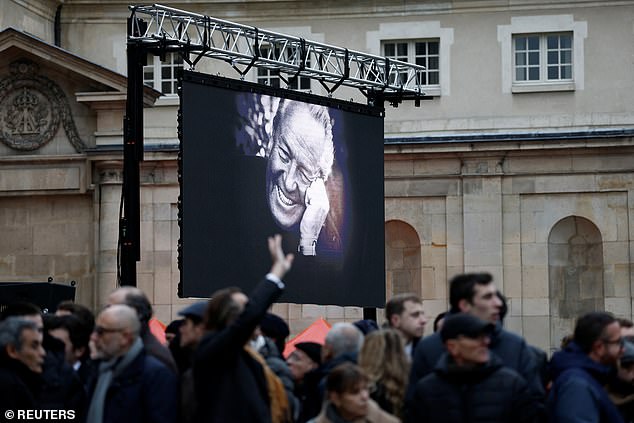 People wait in front of a large screen with a photo of the deceased French far-right figure Jean-Marie Le Pen