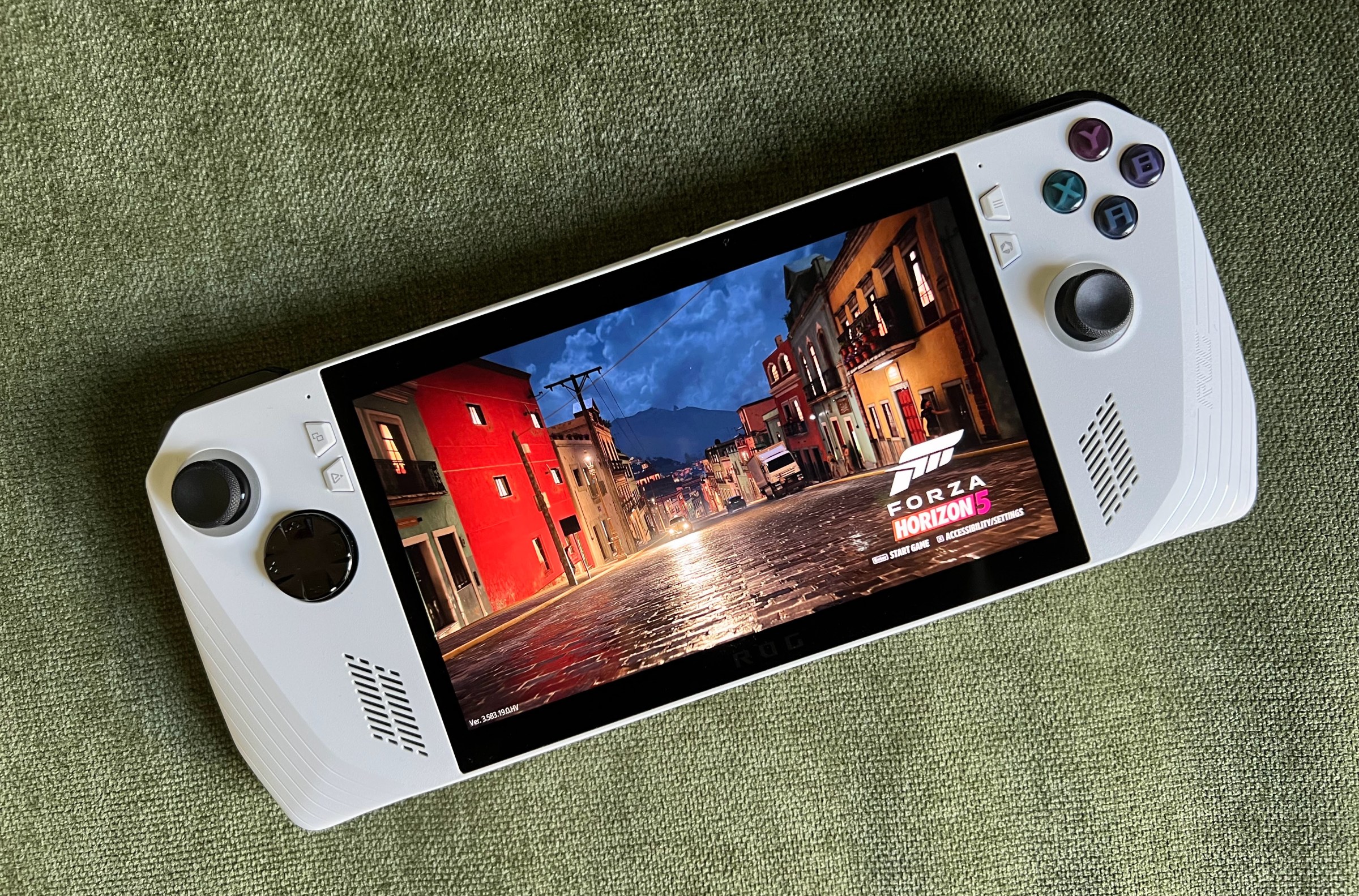 a rainy street scene in Forza Horizon 5 running on an Asus ROG Ally gaming handheld, lying on olive green fabric, photographed from above